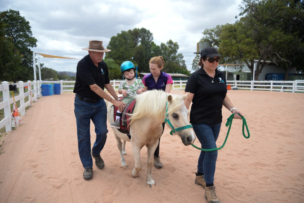 A young boy is on a horse being helped by two volunteers and a therapist.