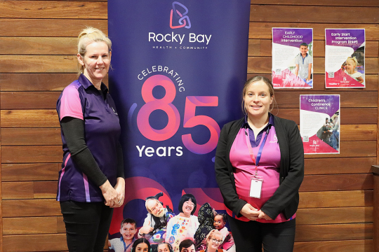 Two staff members stand side by side Rocky Bay's purple 85 Banner.