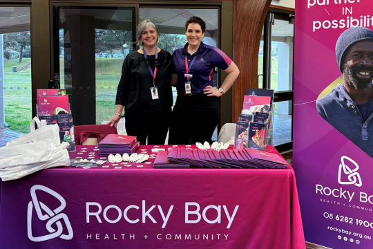 Two cheerful women staffing a vibrant Rocky Bay Health + Community booth at an event. They're standing behind a table draped in a bright pink cloth, showcasing various promotional materials. The booth features the Rocky Bay logo and slogan "partner in possibilities".