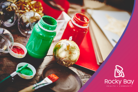 Close-up of Christmas crafting supplies including green and red paint bottles, a gold ornament, paintbrushes, and colorful paper on a wooden surface.