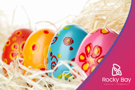 Close-up of four colorfully painted Easter eggs in red, orange, blue, and pink with decorative patterns, nestled in straw. The Rocky Bay Health & Community logo is visible in the bottom right corner against a curved pink border.