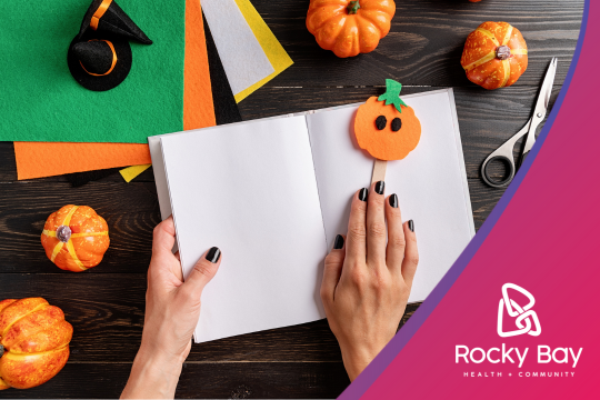 Overhead view of a Halloween crafting scene. Hands with black nail polish hold an open blank notebook. Surrounding items include small orange pumpkins, colorful craft paper, scissors, and a felt pumpkin decoration.