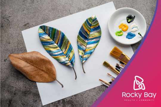 Overhead view of a leaf painting craft. Two colorfully painted leaf shapes in blue, green, and yellow patterns are on a white paper. A natural brown leaf, paint palette with vibrant colors, and various paintbrushes are arranged nearby. The Rocky Bay Health & Community logo is visible in the bottom right corner against a curved pink border.