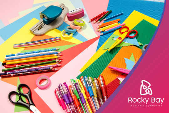 Colorful array of school and craft supplies including pencils, pens, scissors, tape, hole punch, and colored paper, arranged on a vibrant multi-colored background. The Rocky Bay Health & Community logo is visible in the bottom right corner.