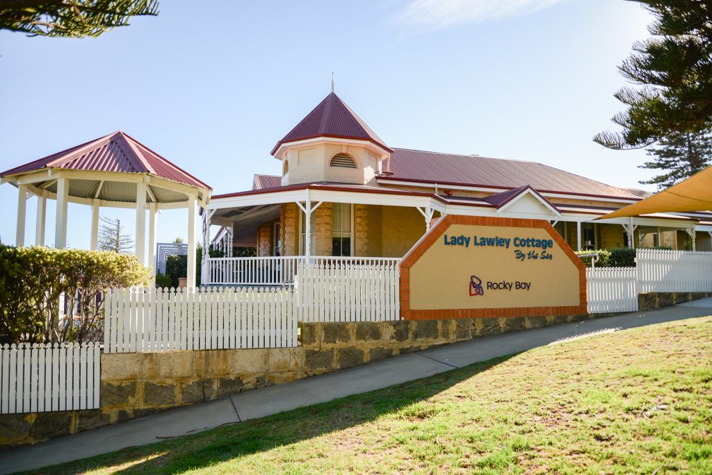 Lady Lawley Cottage by the sea. Yellow building with a burgundy roof and white fence.