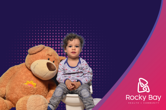 A young child with curly hair sits on what appears to be a stool or pedestal, wearing gray and white patterned pajamas or loungewear. Next to them is a large brown teddy bear. The background features a deep purple color with pink accents and a dotted pattern.